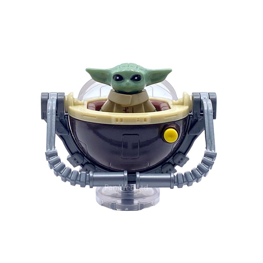 A small green alien toy figure with large black eyes and pointed ears, seated inside a dark bowl-like container with a beige rim and gray mechanical arms, on a clear stand against a white background.